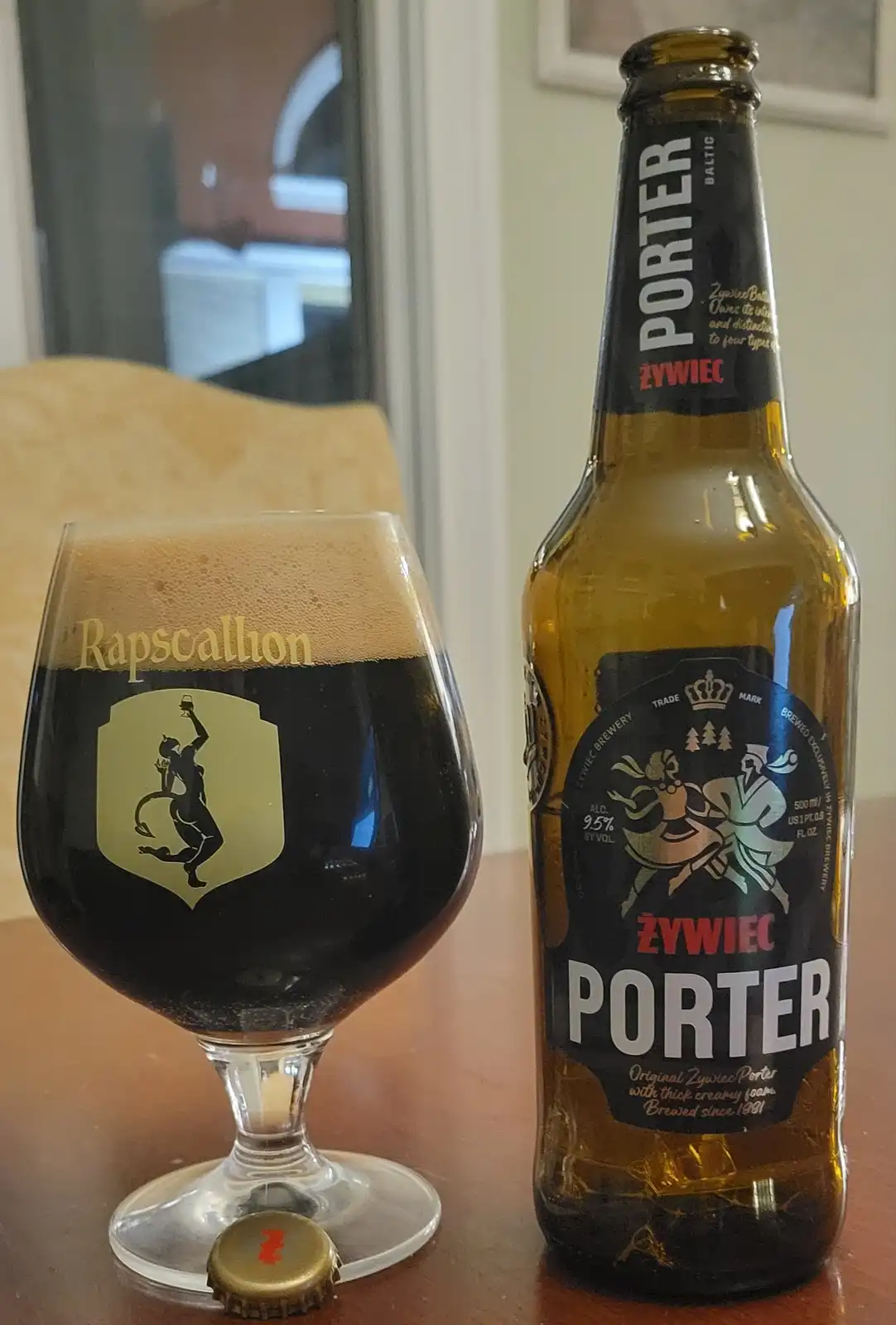 A bottle of Żywiec Porter beer sits next to a snifter glass filled with dark beer and a thick tan head. The glass is branded…