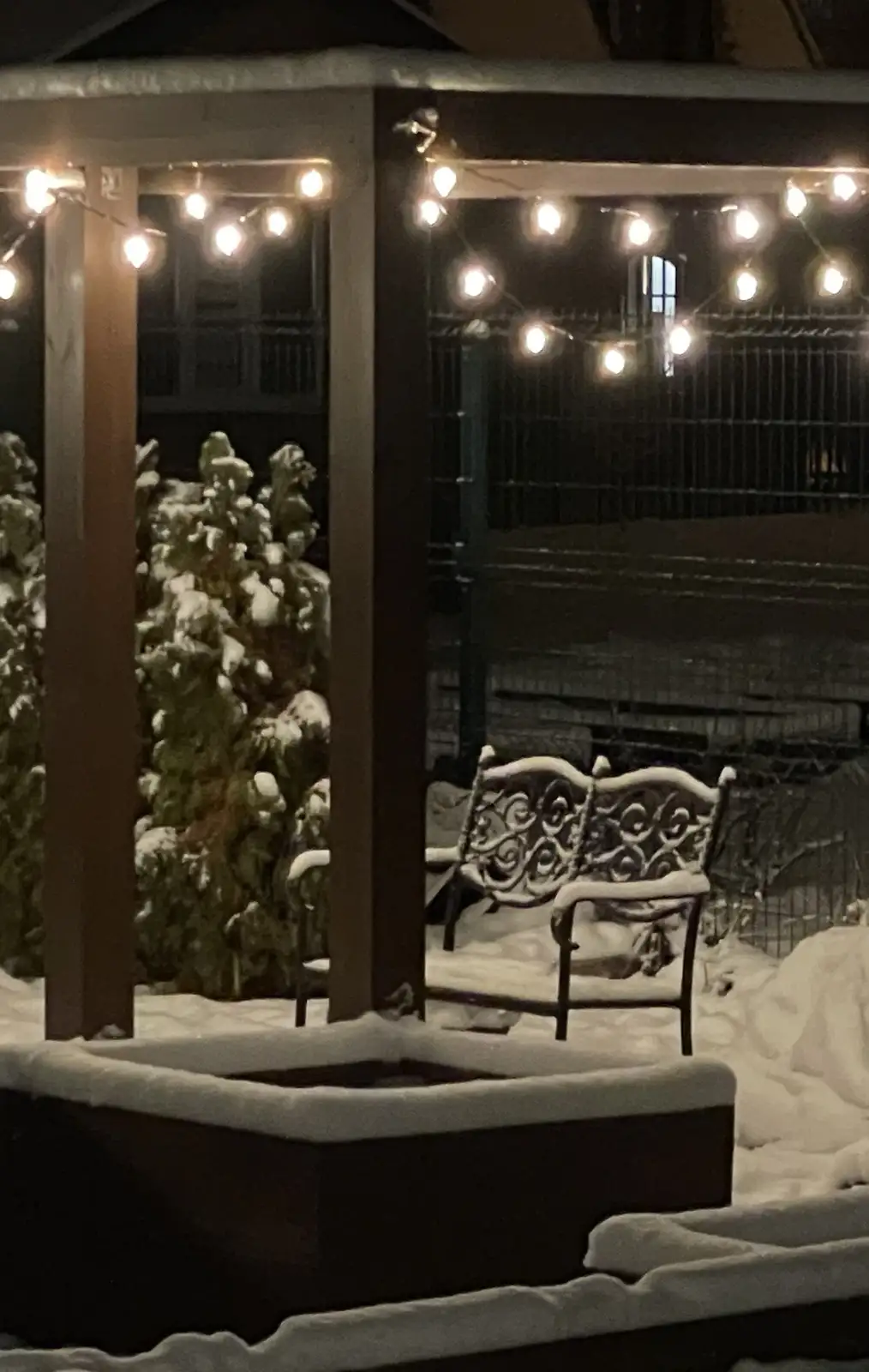 A snowy outdoor patio at night features a snow-covered ornate bench under string lights, with a hot tub also blanketed in…