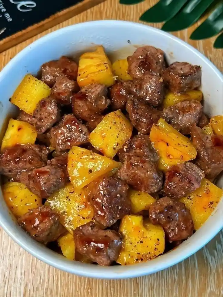 A white bowl filled with bite-sized pieces of cooked steak mixed with chunks of yellow pineapple, seasoned with black pepper…