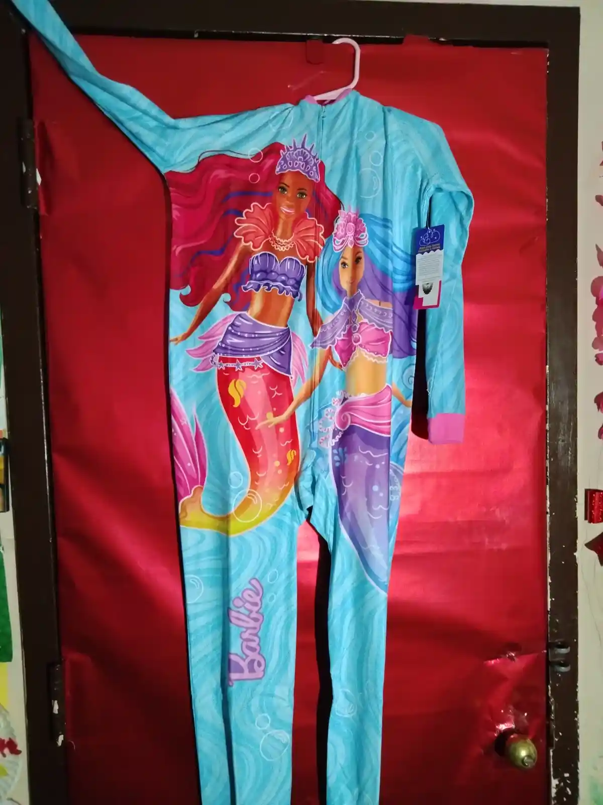 A light blue, full-body children's onesie with a Barbie mermaid design hangs on a pink hanger against a red door. The front…