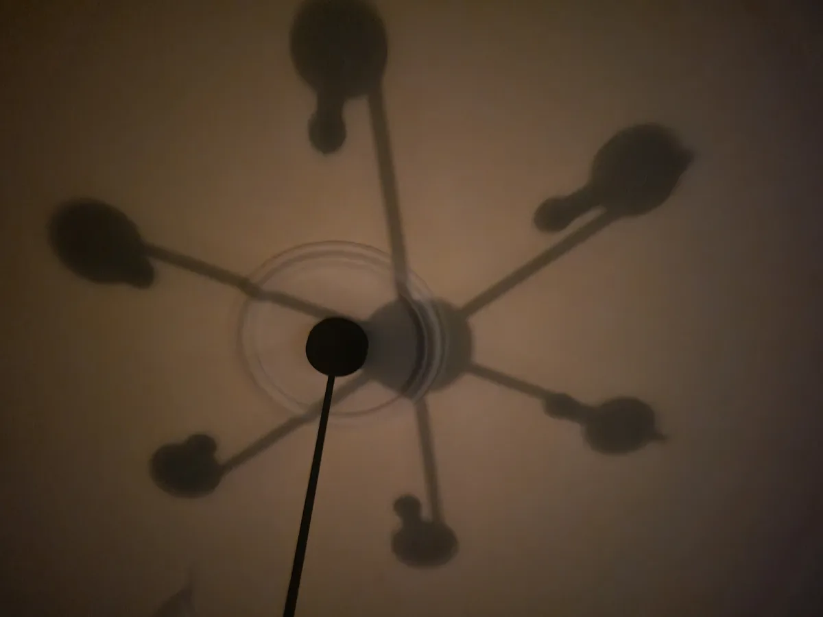 A dimly lit ceiling shows the shadow of a modern chandelier with multiple arms and bulbs, casting a dark, somewhat…