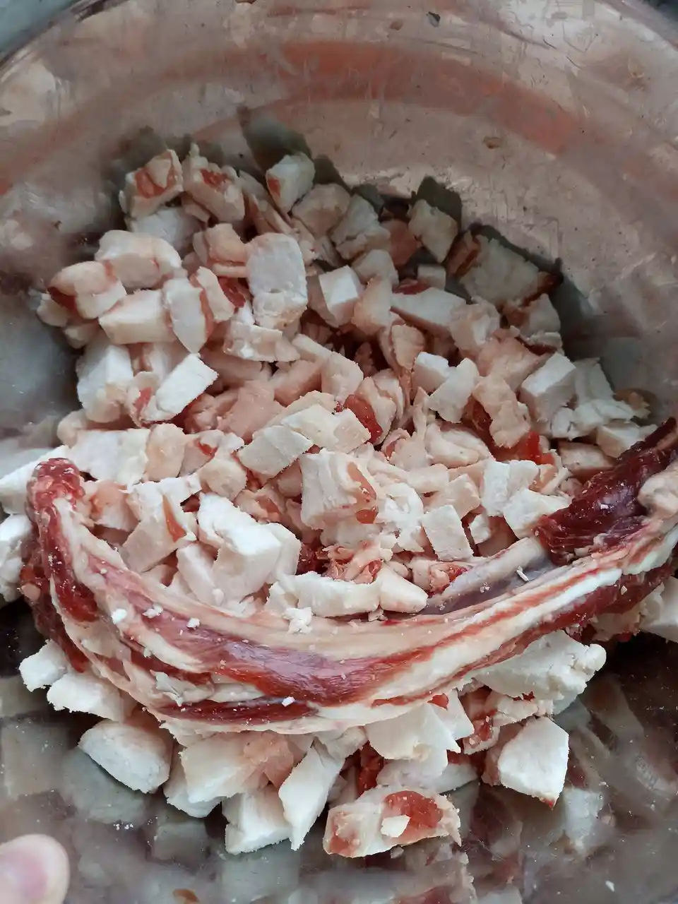 A glass bowl contains chopped raw chicken pieces mixed with a single, unbroken strip of bacon resting on top. The text…