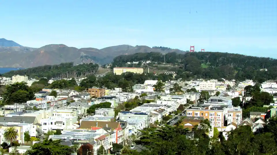 A scenic view of a hilly cityscape with colorful houses, trees, and distant mountains under a clear blue sky. The iconic red…