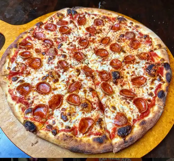 A close-up photo of a pepperoni pizza on a wooden serving board, with visibly charred and blackened spots on the crust and…