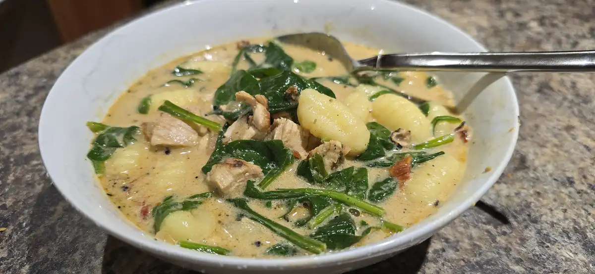 A close-up photo shows a white bowl filled with creamy gnocchi soup containing spinach, chicken pieces, and gnocchi, resting…