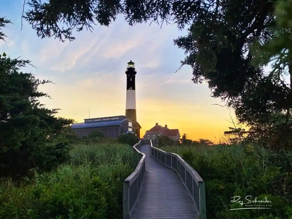 A scenic photo of a lighthouse at sunset, viewed through trees, with a wooden boardwalk leading toward it. The image has…