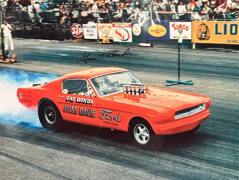 A vintage red Ford Mustang drag race car, driven by Russ Davis, is shown performing a burnout on a racetrack, kicking up…