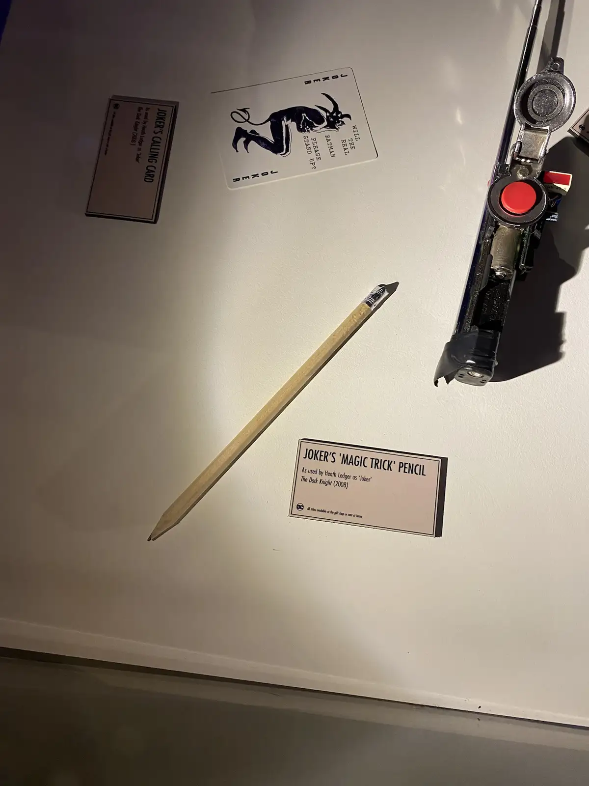 A museum display shows a simple wooden pencil labeled 'Joker's Magic Trick Pencil' from The Dark Knight (2008), alongside a…
