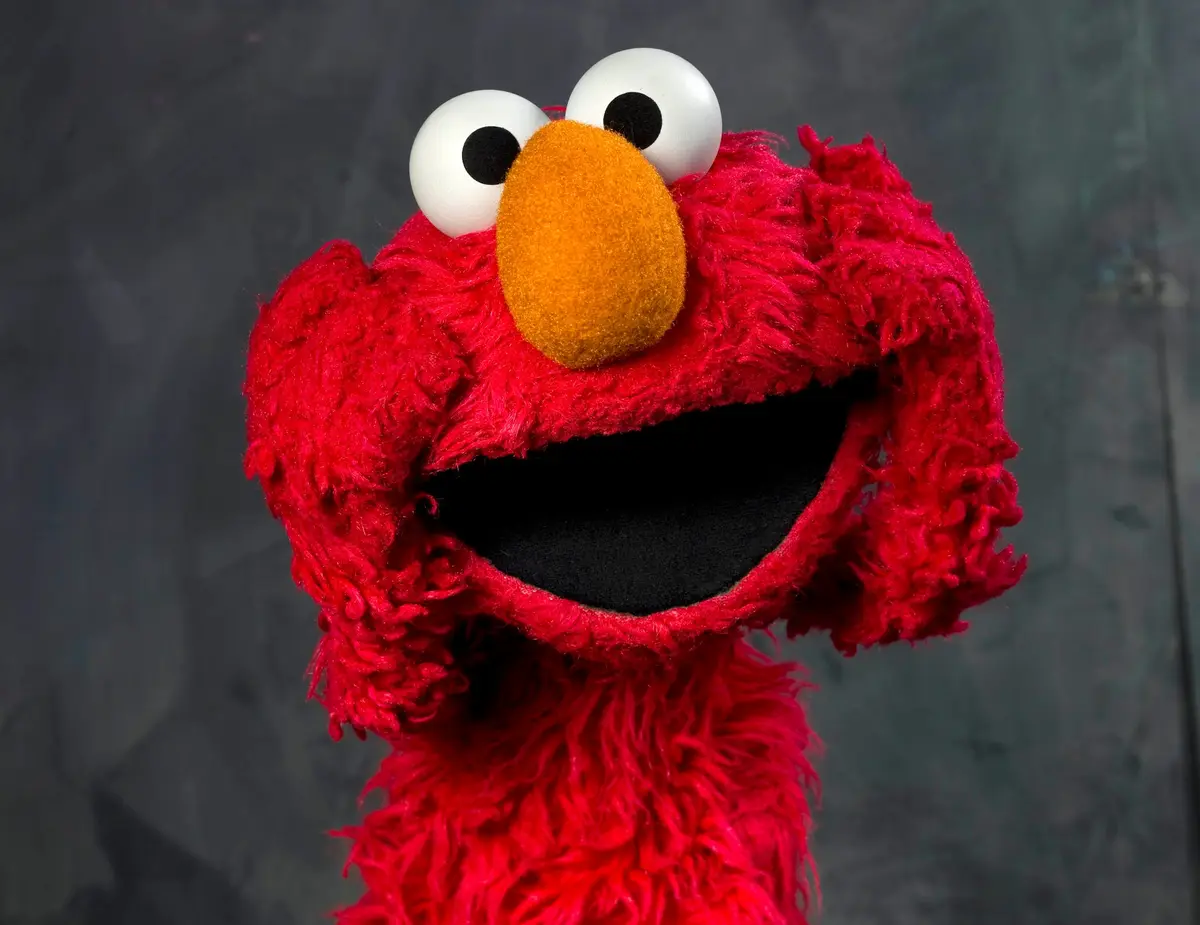 A close-up of the red, furry Sesame Street character Elmo with his mouth wide open in a joyful expression against a dark…