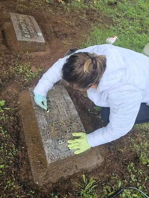 A person wearing a white puffer jacket and gloves is kneeling in a muddy cemetery, using a small brush to clean the engraved…