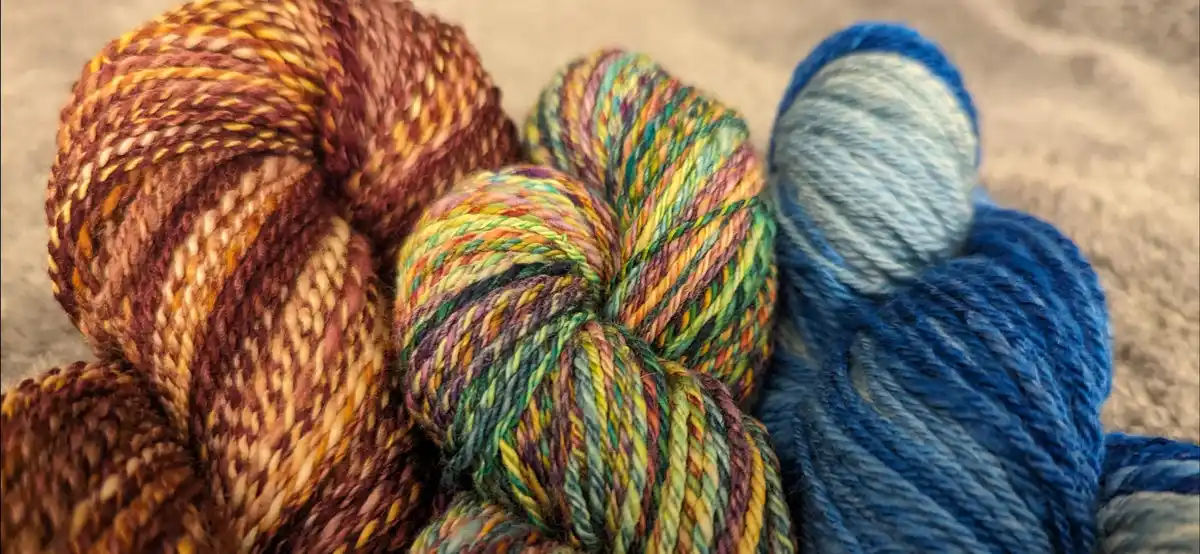 Three skeins of colorful, variegated yarn are arranged side-by-side on a textured beige surface. The left skein is earthy…