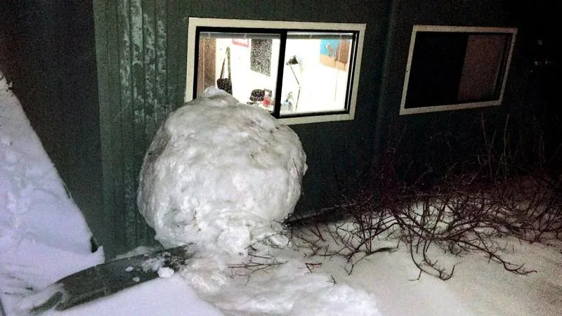 A large, roughly spherical snowball sits on the snowy ground outside a dark building at night, partially obscuring a window…