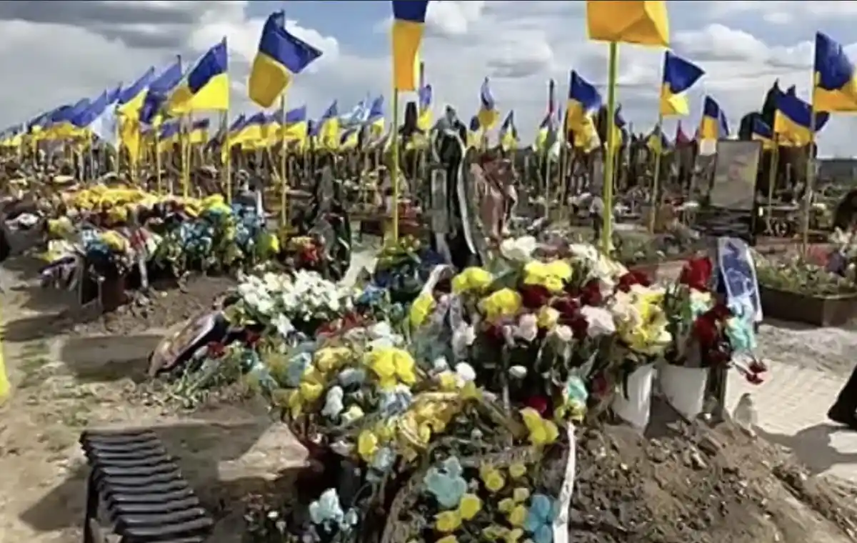 A wide shot of a cemetery filled with freshly dug graves, each adorned with abundant floral arrangements and numerous…