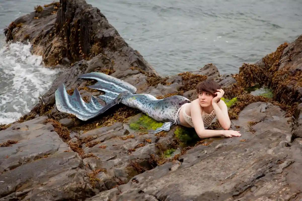 A person with short brown hair lies on wet, mossy rocks at the ocean's edge, wearing a mermaid tail costume with blue and…