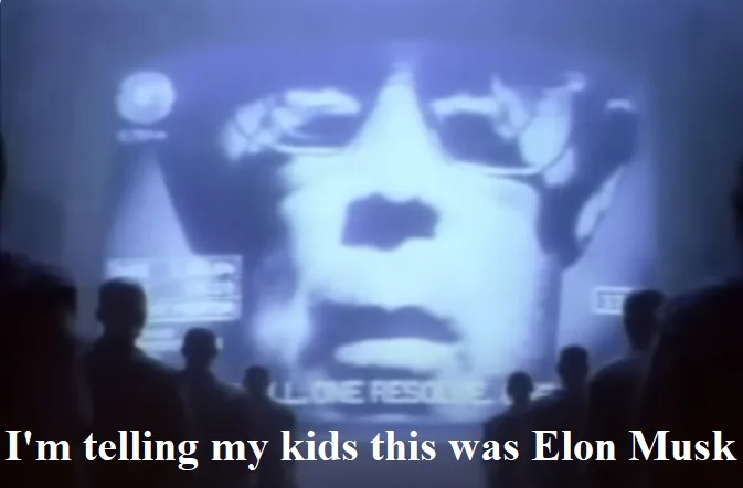 A grainy, blue-tinted image shows a large projected face of a man wearing glasses, with silhouettes of people in the…