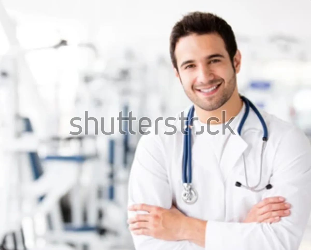 A smiling man in a white lab coat with a stethoscope around his neck stands with arms crossed in a blurry medical setting…