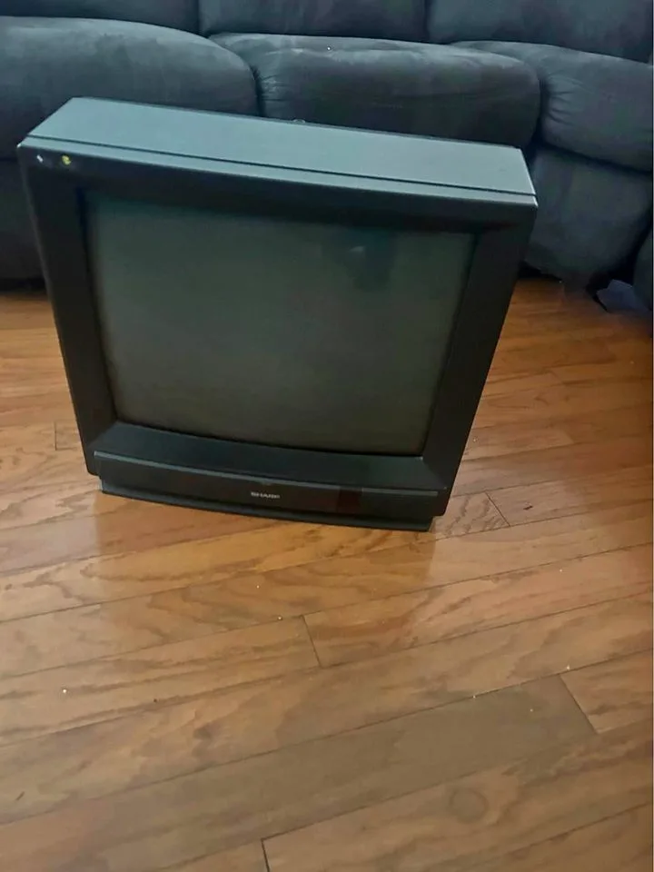 A black, boxy, old-style CRT television sits upright on a wooden floor, with a dark gray couch visible behind it. The TV…