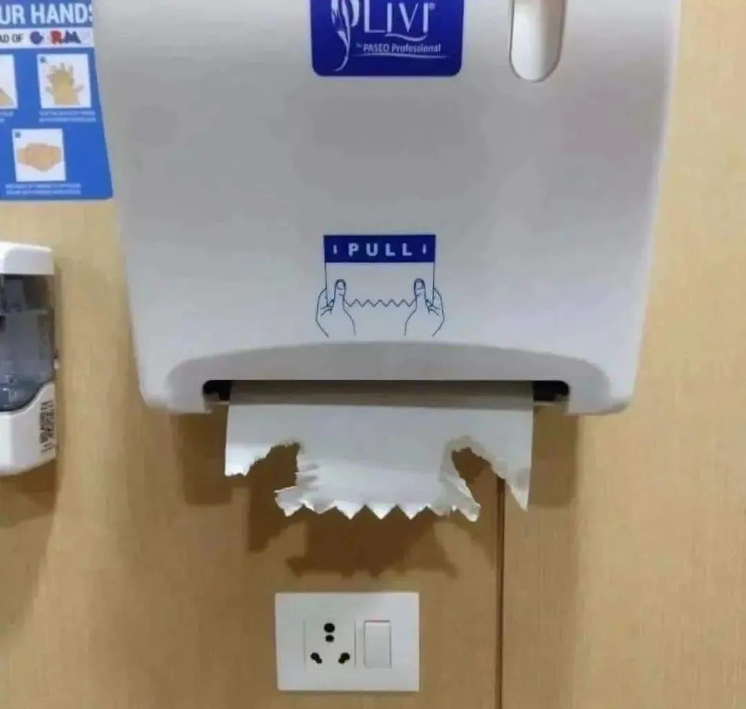 A white wall-mounted paper towel dispenser with a blue 'PULL' instruction sticker showing hands tearing paper. The paper…