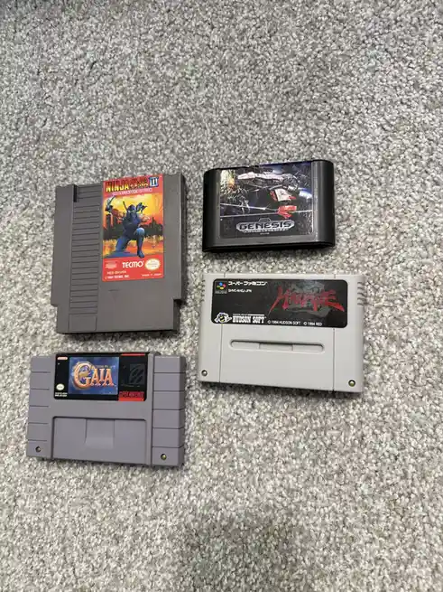 Four retro video game cartridges — Ninja Gaiden III, a Genesis game, Macne, and Gaia — are laid out on a gray carpet. The…
