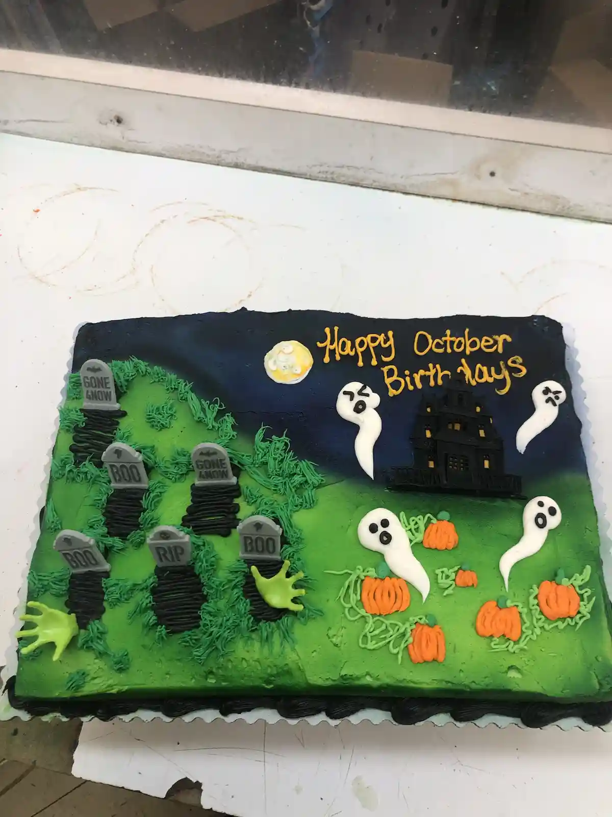 A rectangular Halloween-themed birthday cake decorated with green frosting resembling grass, black frosting for a night sky…