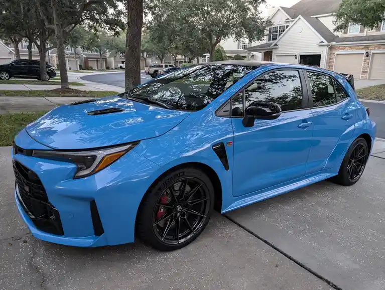 a bright blue toyota corolla gr sport is parked on a residential driveway, featuring black accents, black wheels with red…