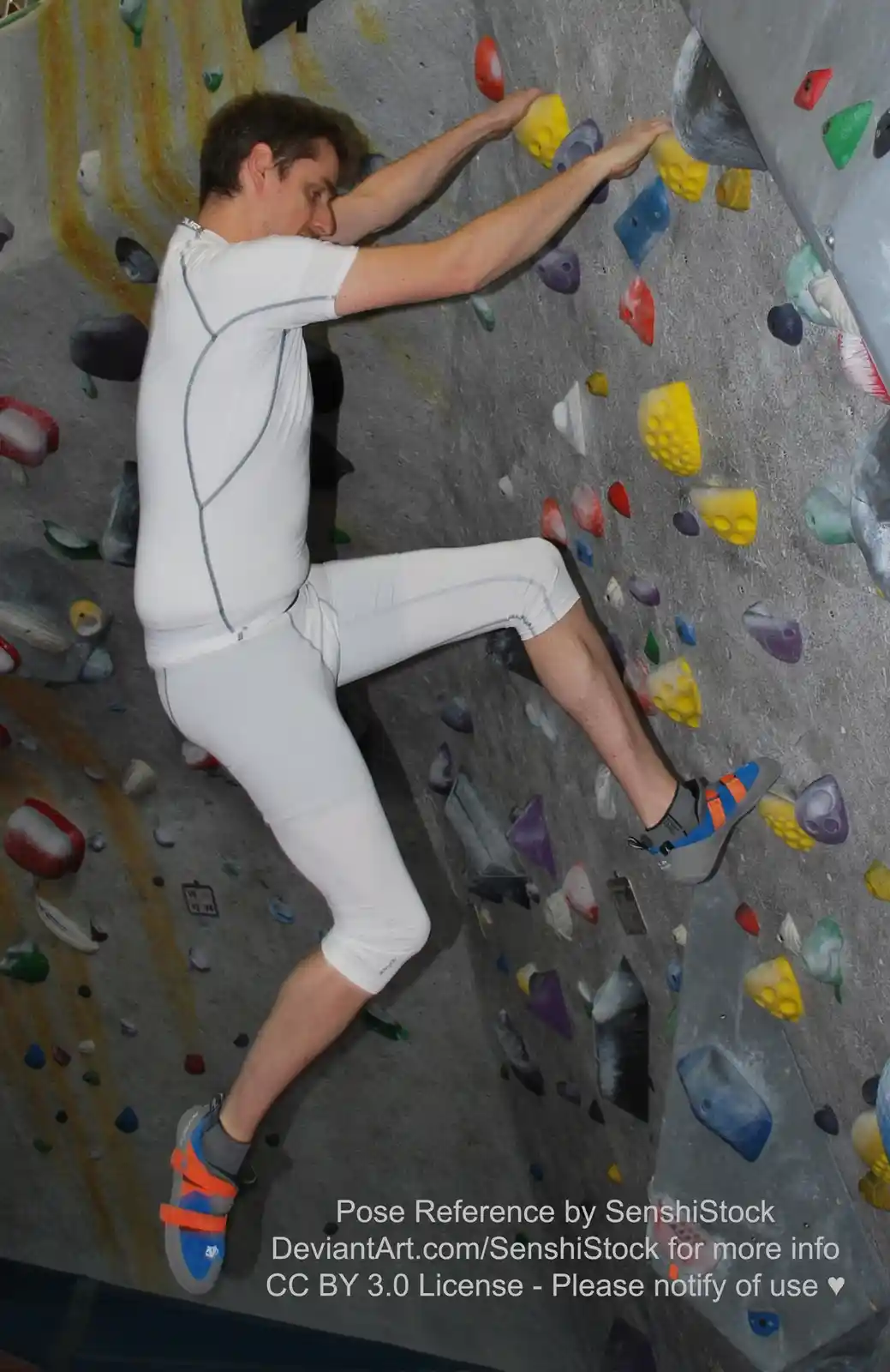 A man in white athletic wear is climbing an indoor rock wall, gripping colorful handholds with both hands and one foot while…