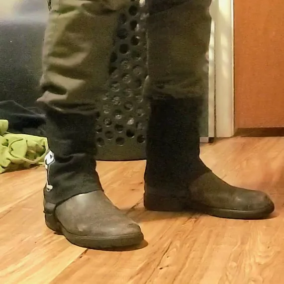 A person wearing dark pants and black boots stands on a wooden floor, with one boot visibly pulled up over the pant leg…