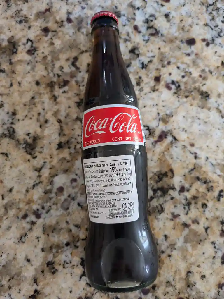 A glass Coca-Cola bottle with a red cap and label sits on a speckled granite countertop, showing its nutrition facts label…