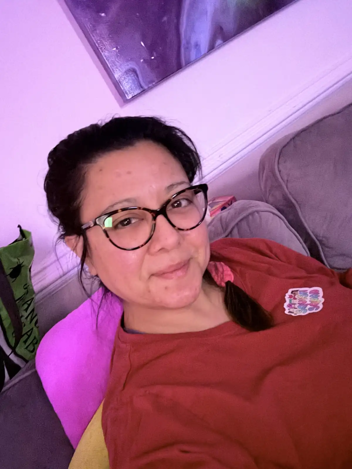 A woman with dark hair pulled back and wearing tortoiseshell glasses lies on a couch under purple ambient lighting. She…