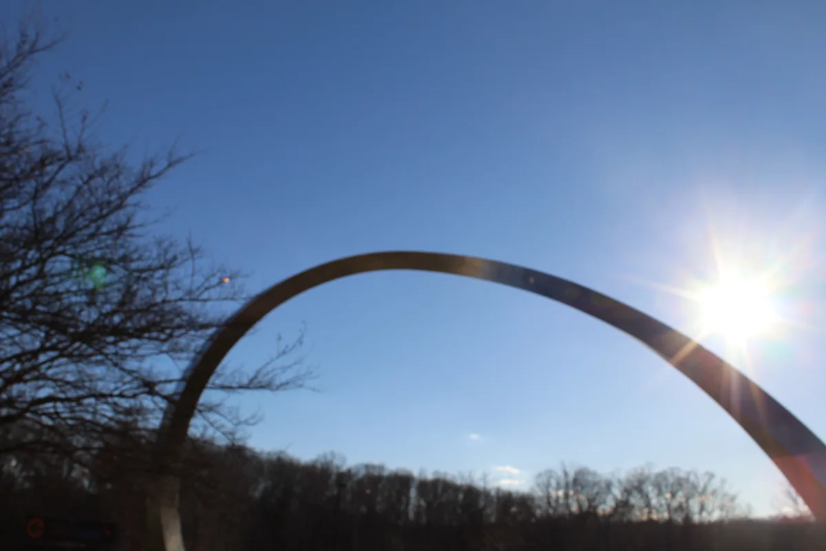 A low-angle photo shows the Gateway Arch in St. Louis against a clear blue sky, with the sun shining brightly through the…
