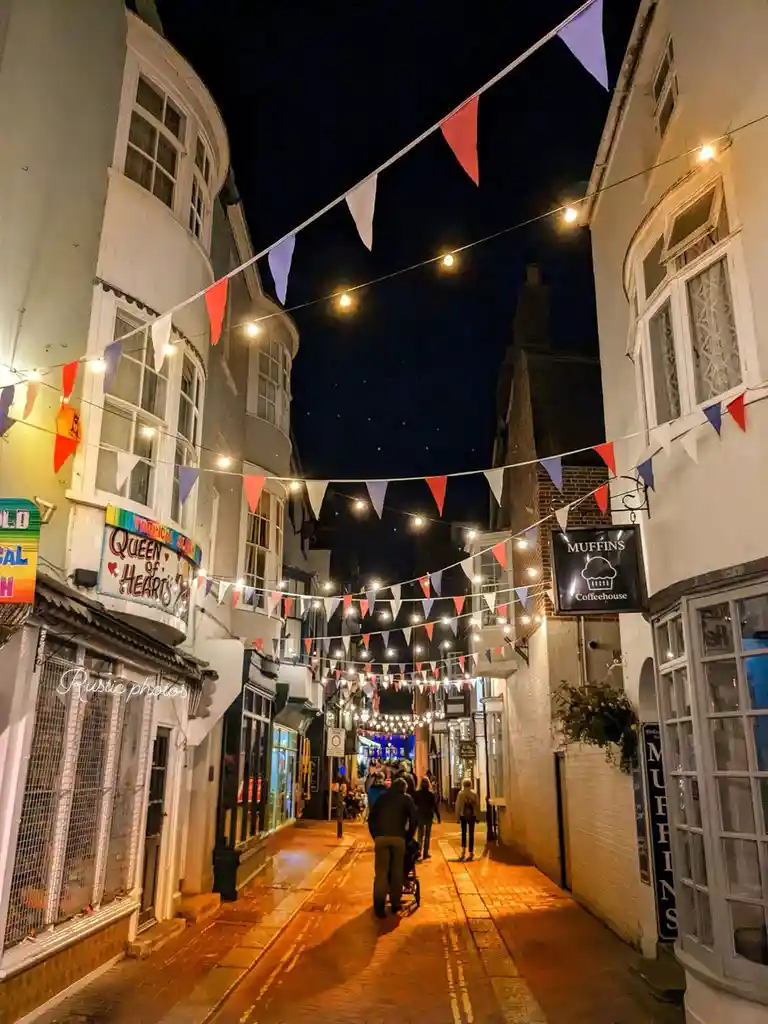 A nighttime photo of a narrow, charming street lined with historic buildings, illuminated by string lights and red, white…