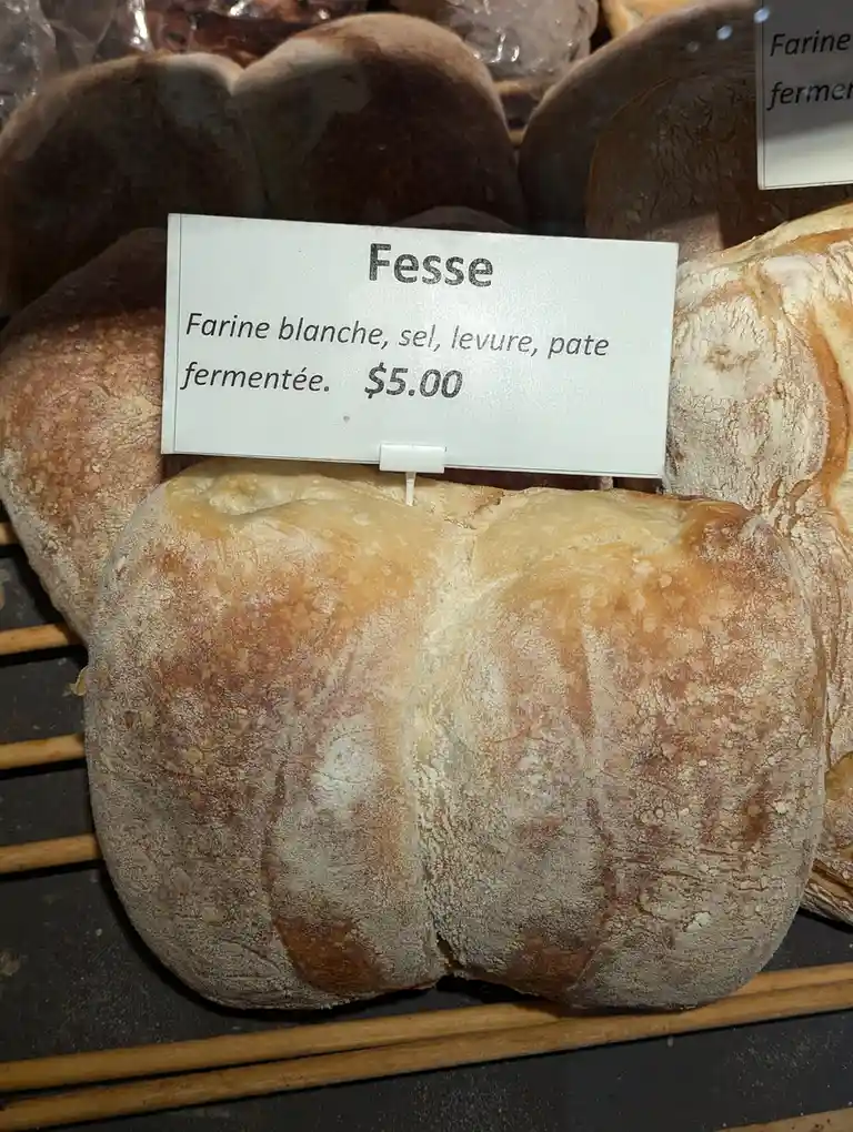 A loaf of bread with a distinctively rounded, two-buttock shape sits on a bakery display rack, labeled 'Fesse' with…