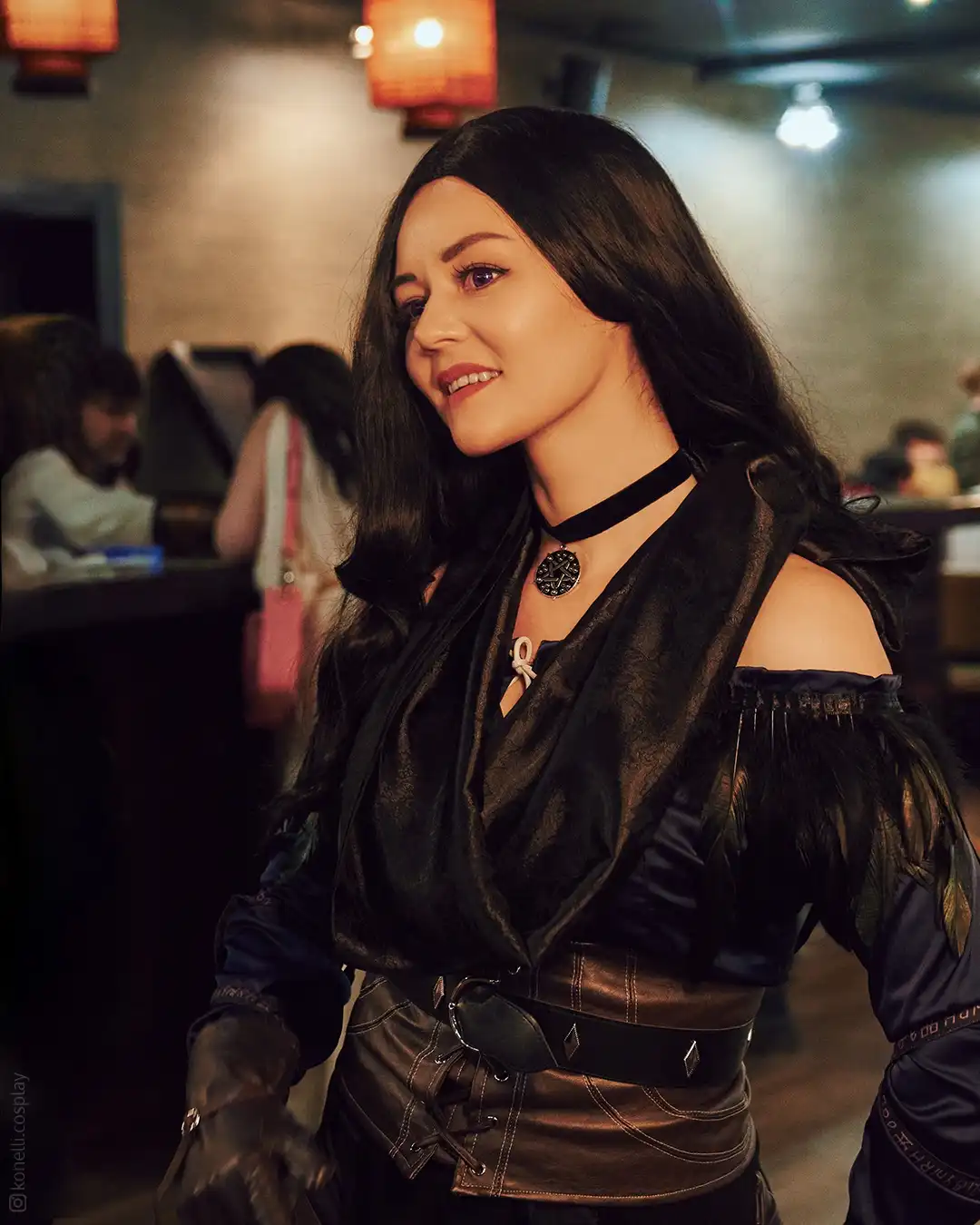 A woman in a dark, gothic-style costume with long black hair, a choker, and leather corset smiles confidently at the camera…