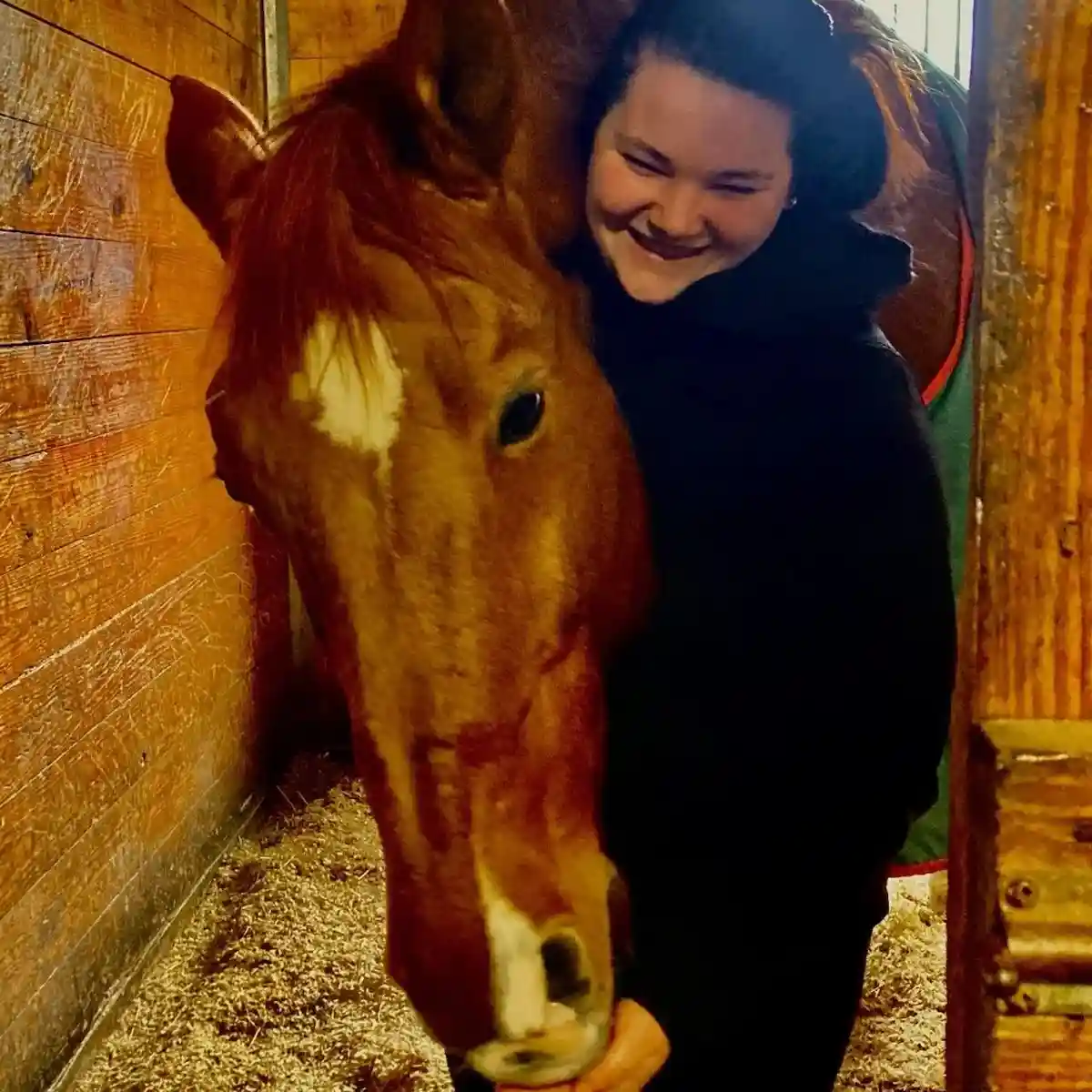 A person in a black hoodie is smiling broadly while standing close to a brown horse in a wooden stable, with the horse's…