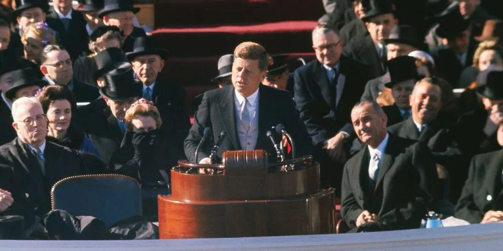 A historical photograph shows President John F. Kennedy standing at a wooden podium during an outdoor event, surrounded by…