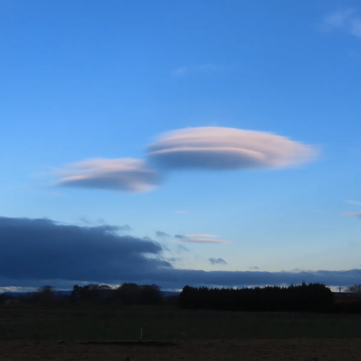 A landscape photo shows a clear blue sky with a prominent, smooth, lens-shaped cloud resembling a UFO hovering above a dark…