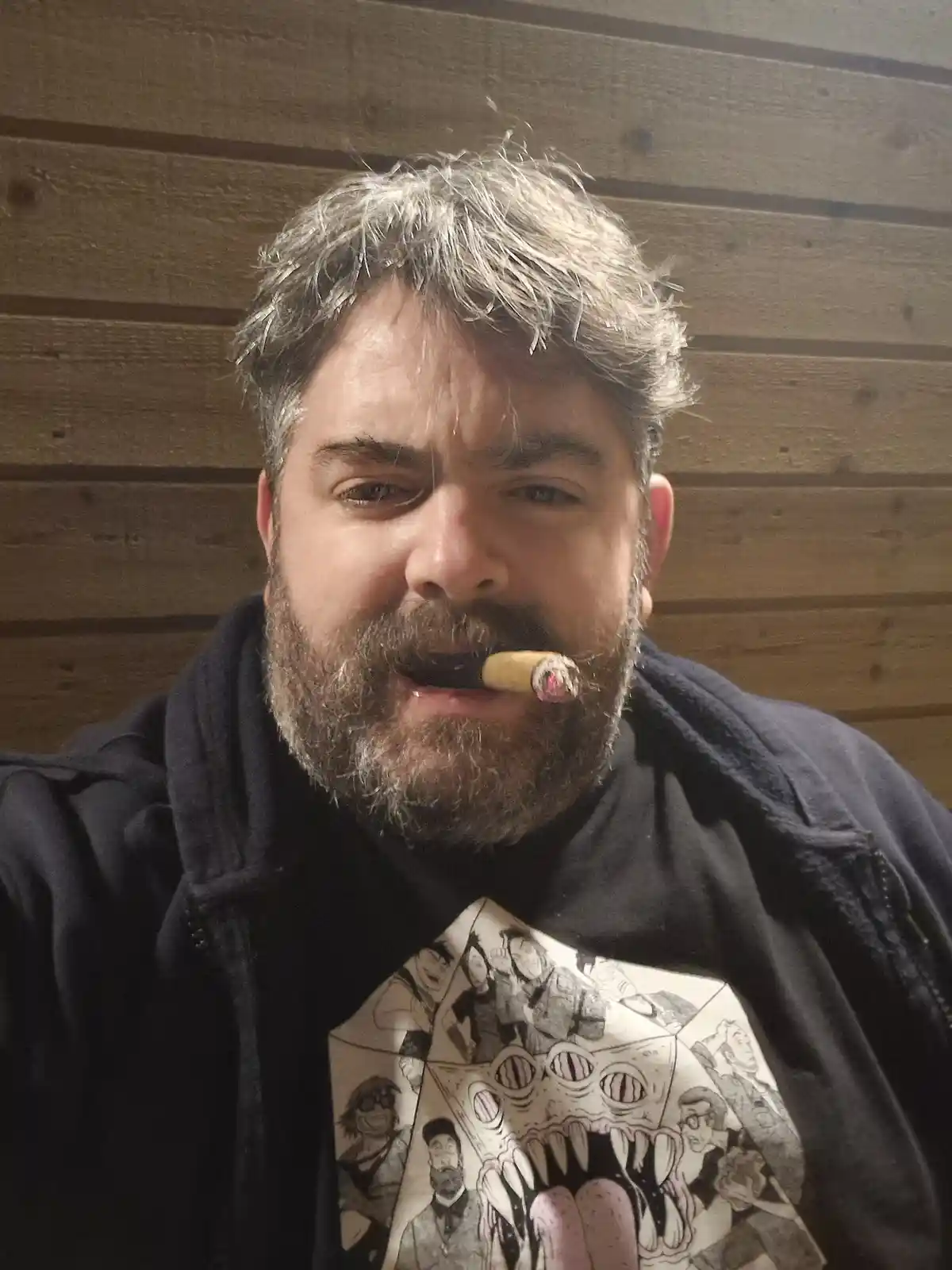 A man with a beard and gray hair, wearing a dark hoodie over a graphic t-shirt, holds a lit cigar in his mouth. He stares…
