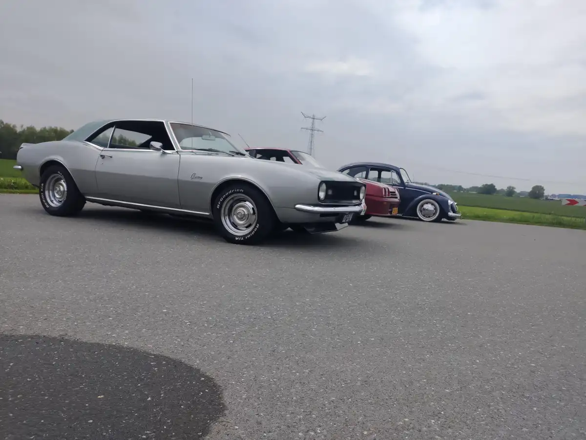 Three classic cars are parked side-by-side on an asphalt road: a silver Chevrolet Camaro in the foreground, a red and blue…