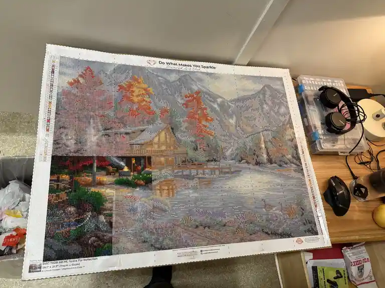 A partially completed diamond painting of a cabin in autumn with mountains, showing a large section of the image still blank…