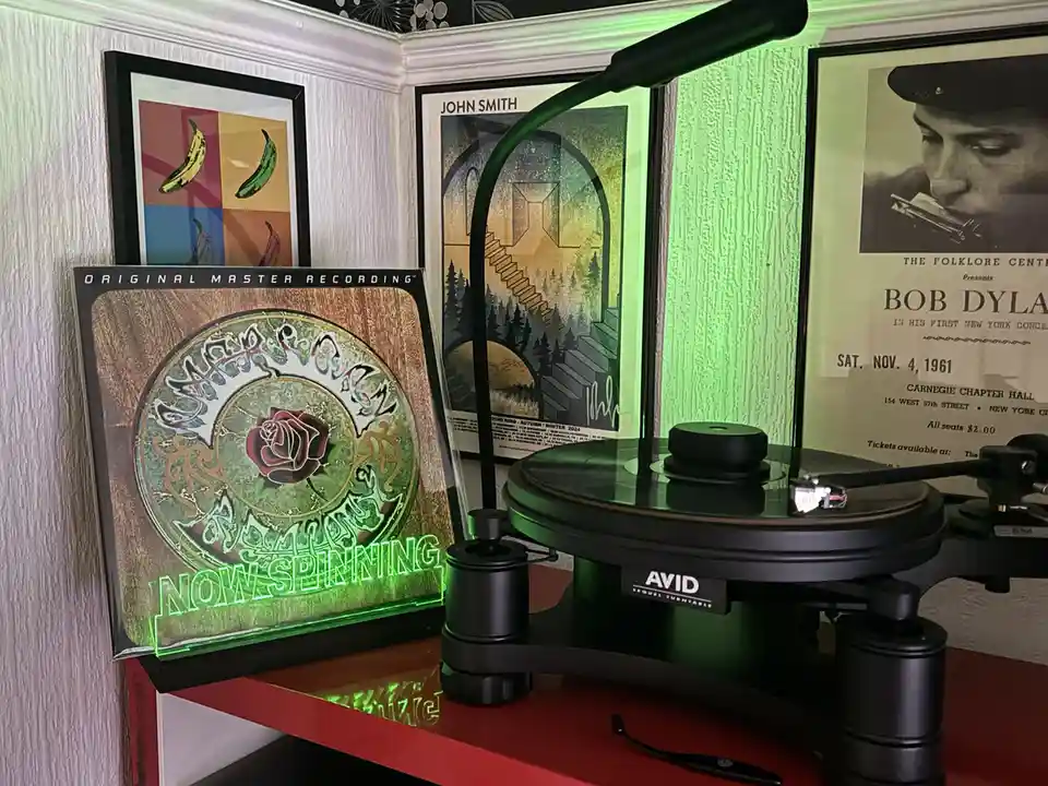 A vinyl record player with a glowing green 'NOW SPINNING' sign next to a framed album cover. The background features framed…
