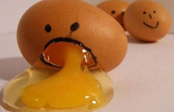 A cracked brown egg with a sad face drawn on it is leaking yellow yolk onto a white surface, while two other eggs with…