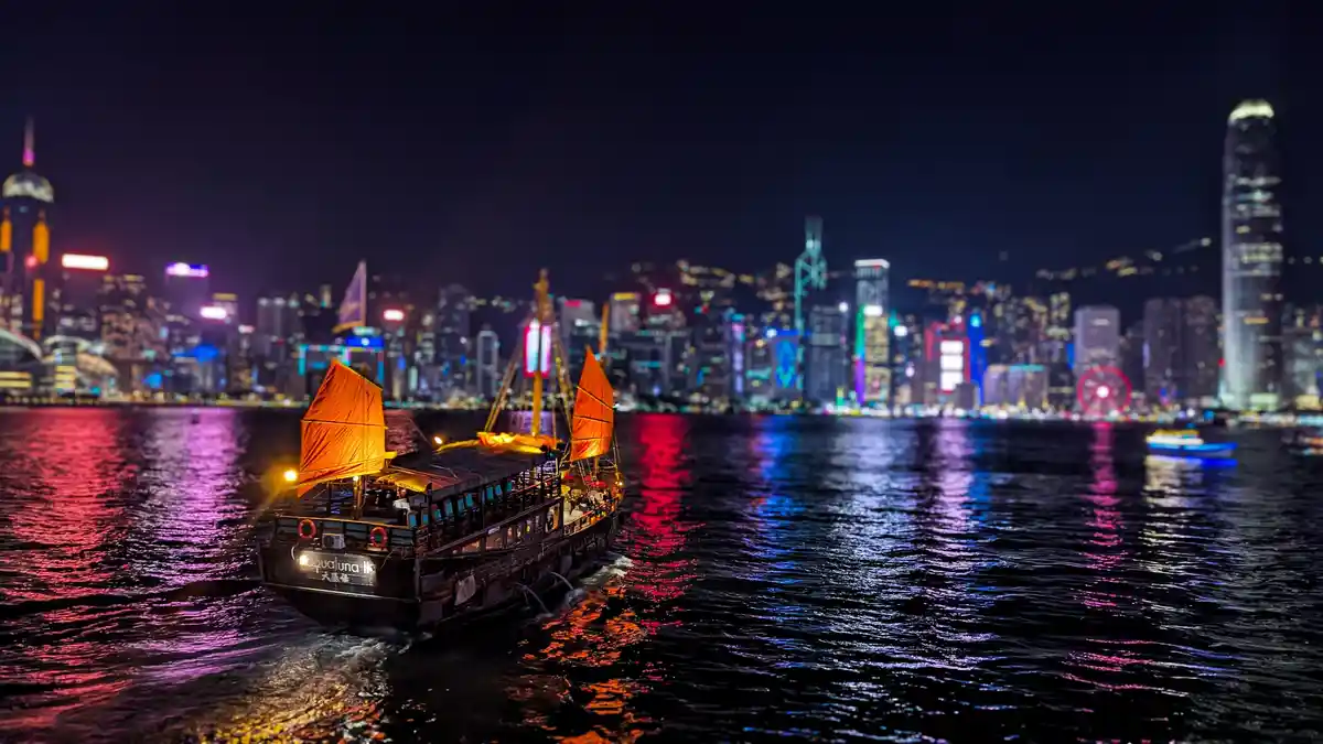 A traditional Chinese junk boat with orange sails sails on a dark waterway at night, leaving a wake. The background is a…