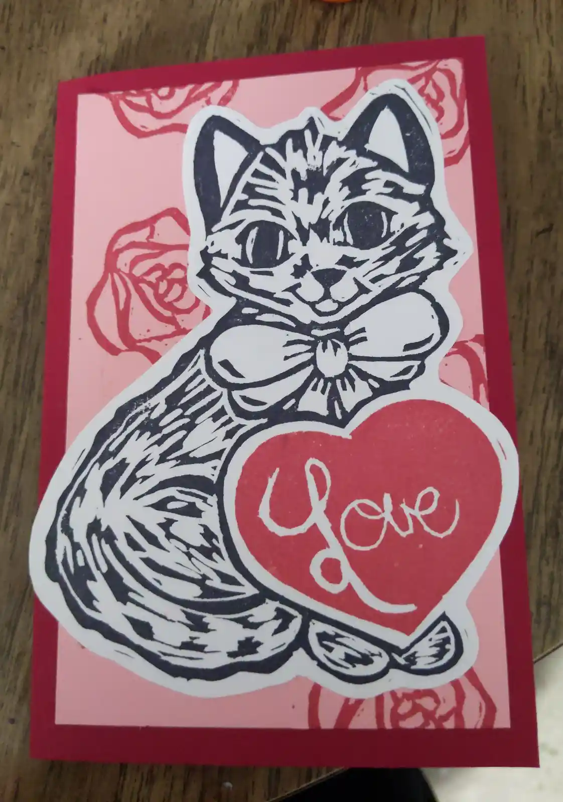 A handmade Valentine's card features a black-and-white linocut-style illustration of a cat wearing a bowtie, holding a red…