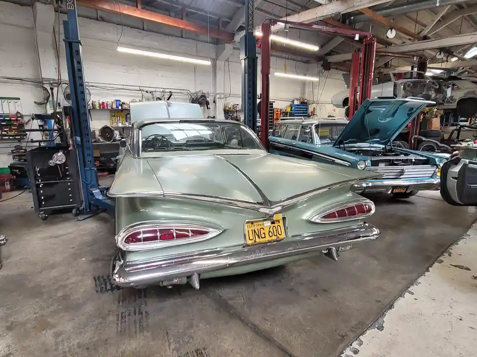 A light green 1950s classic car with a California license plate reading 'UNG 600' is parked in a cluttered auto repair…