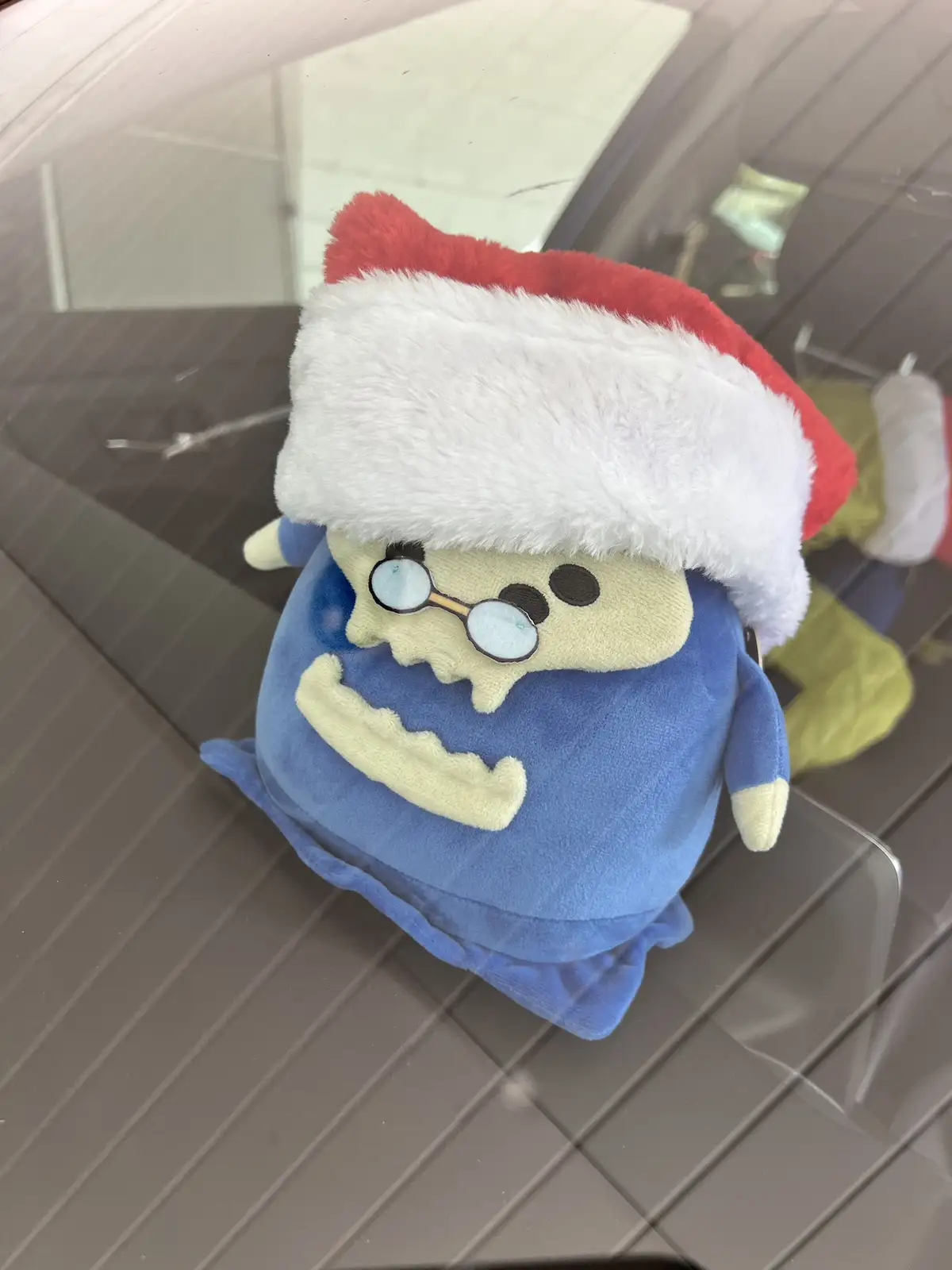 A blue plush toy resembling a cartoonish, skeletal character with glasses and a wide grin, wearing a red and white Santa…