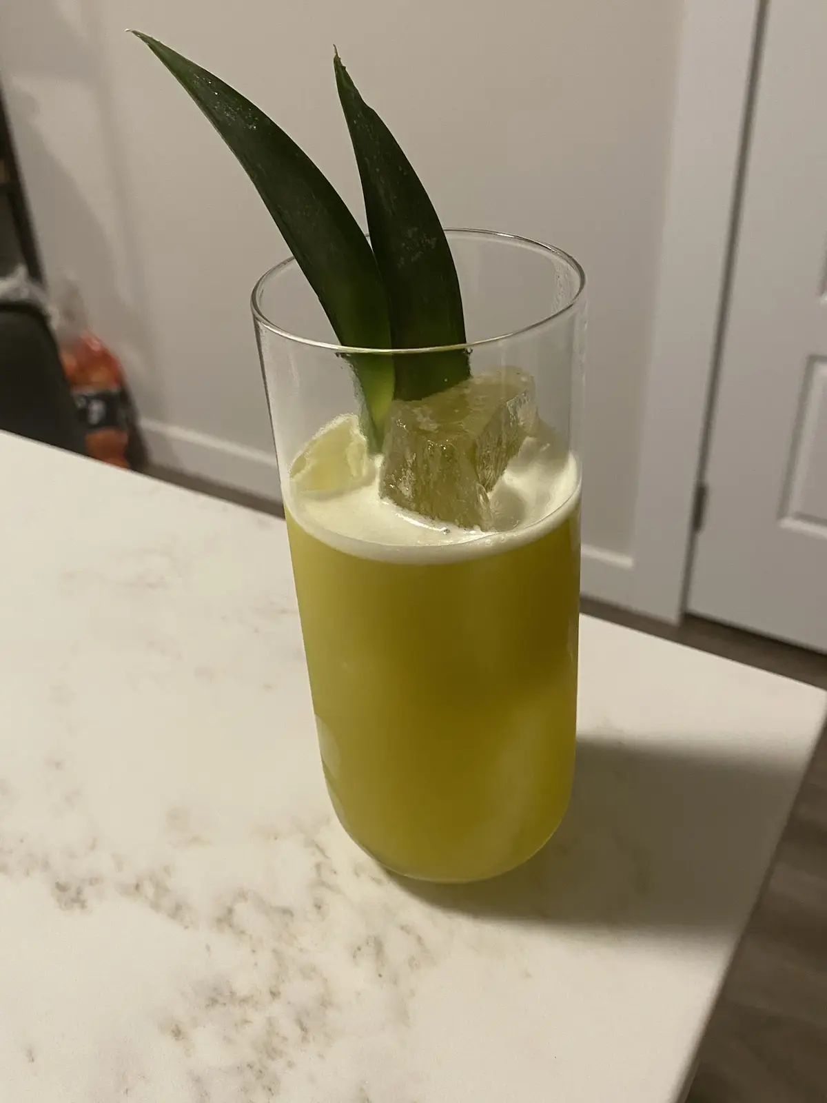 A tall clear glass filled with a yellow liquid and a large ice cube sits on a white marble countertop, garnished with two…