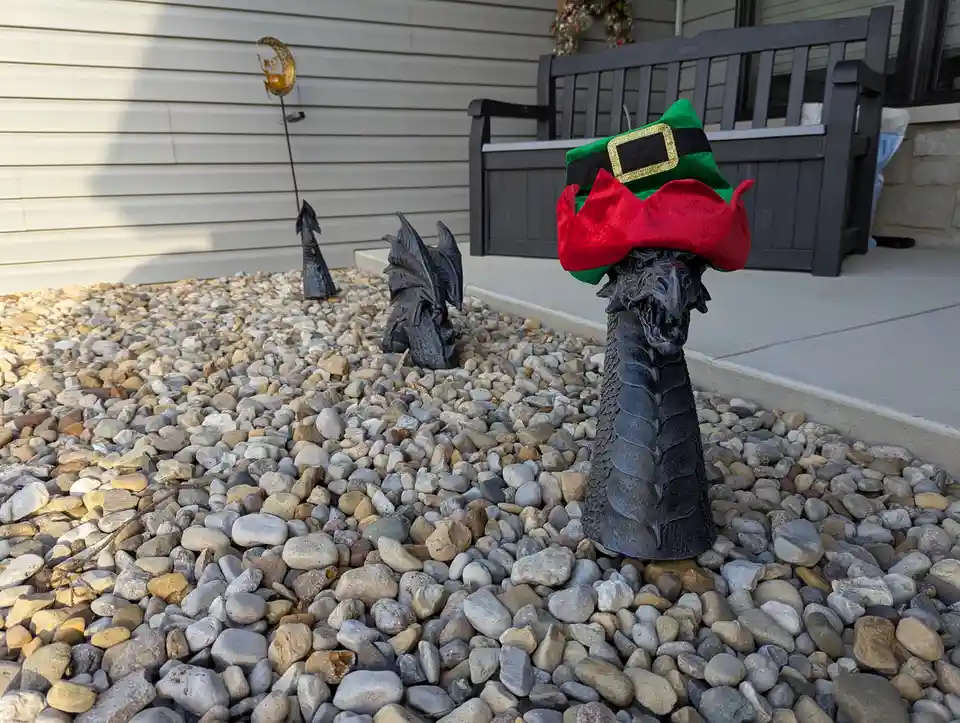 A black plastic dragon garden ornament stands in a bed of pebbles, wearing a red and green elf hat with a gold buckle…