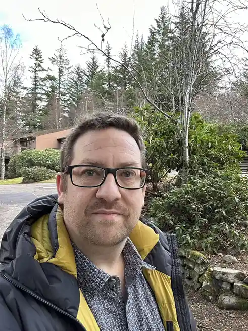 A man with glasses and a black-and-yellow jacket looks at the camera with a neutral expression, standing outdoors near trees…