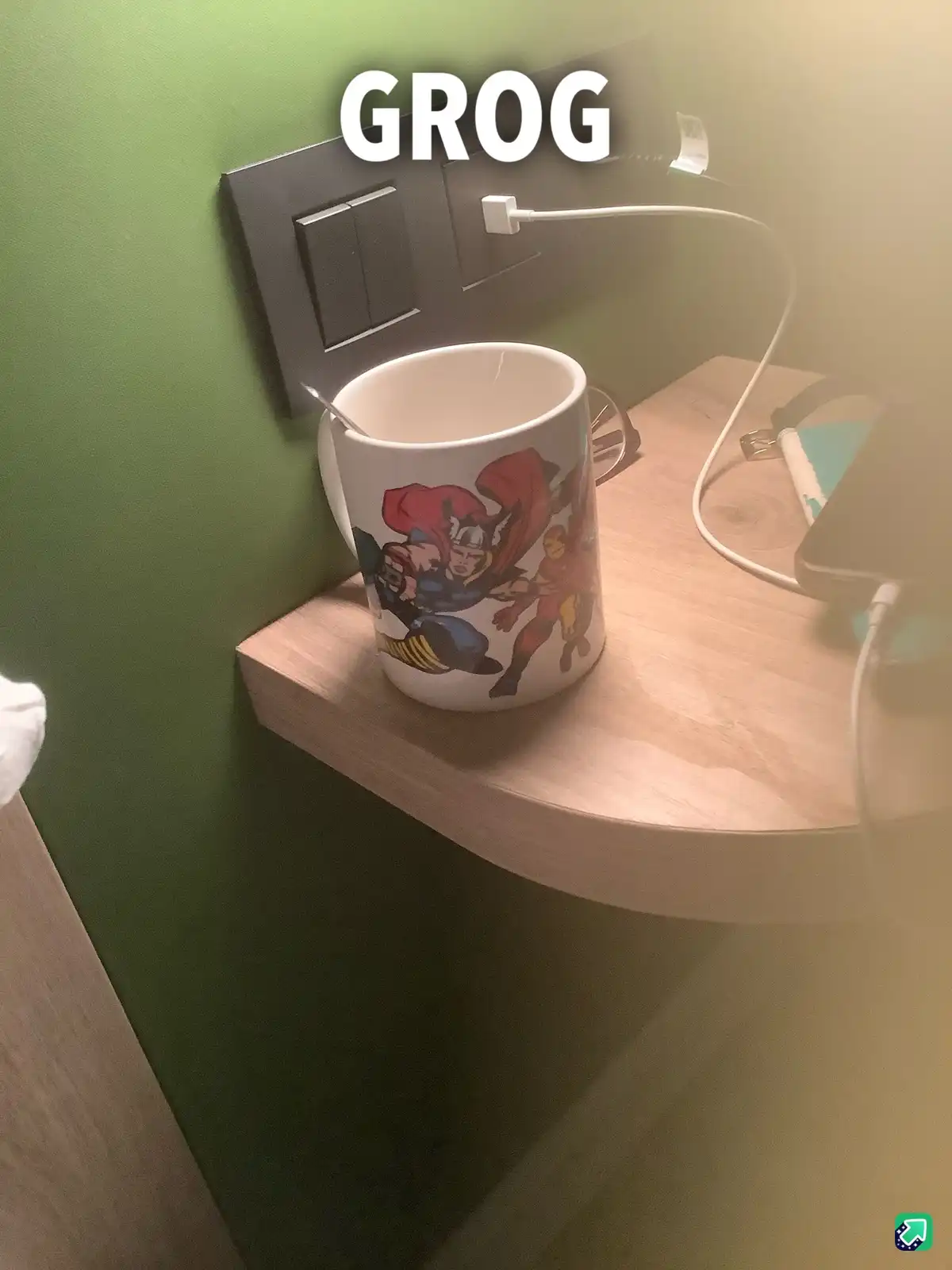 A white ceramic mug with comic book superheroes printed on it sits on a light wooden bedside table next to a green wall. A…