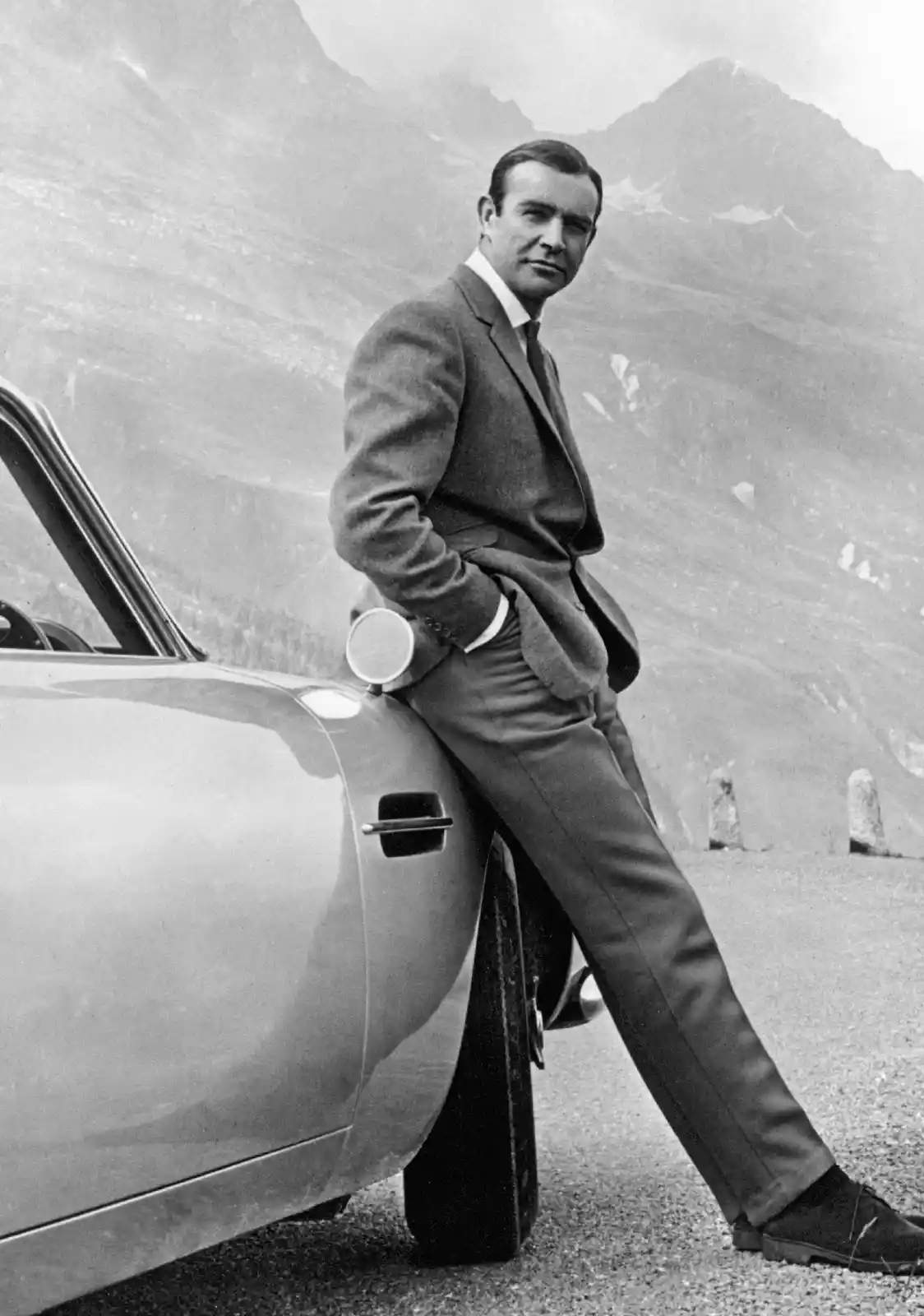 A black-and-white photo shows Sean Connery leaning against a vintage sports car with mountains in the background. He wears a…